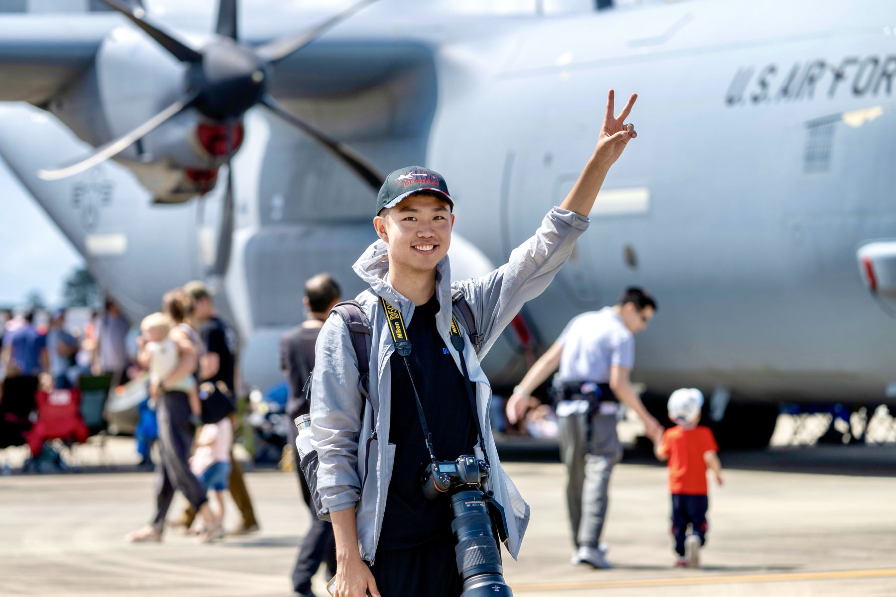 Thomas waving in front of a U.S. Air Force propeller transport aircraft at an air show, camera in hand