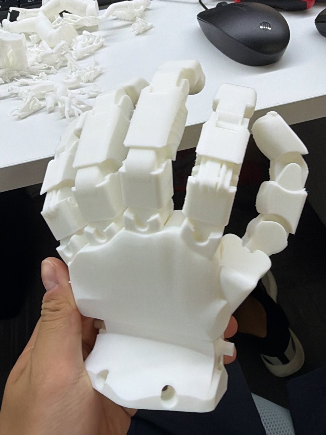 A white 3D-printed robot hand held in one hand above a desk