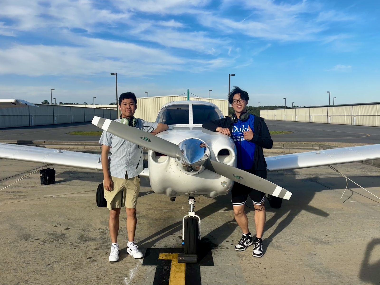 Two students standing on either side of the propeller of a small white aircraft on the ramp